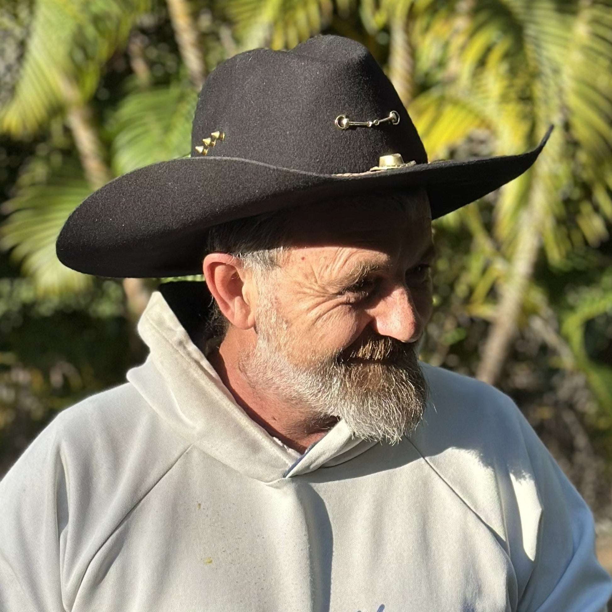 Man wearing a black cowboy hat with decorative elements, standing outdoors with greenery in the background.